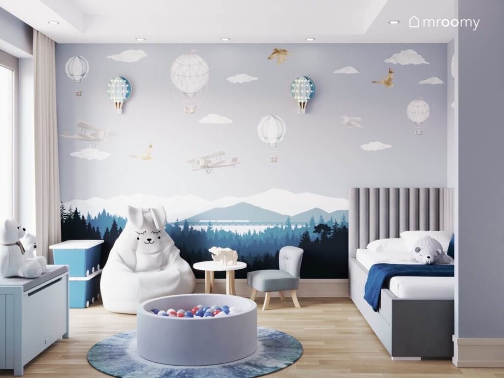 Szaroniebieska ściana w pokoju chłopca na niej górska panorama naklejki samoloty balony i chmurki oraz urocze lampki balony a pod spodem pufa królik oraz szare łóżko