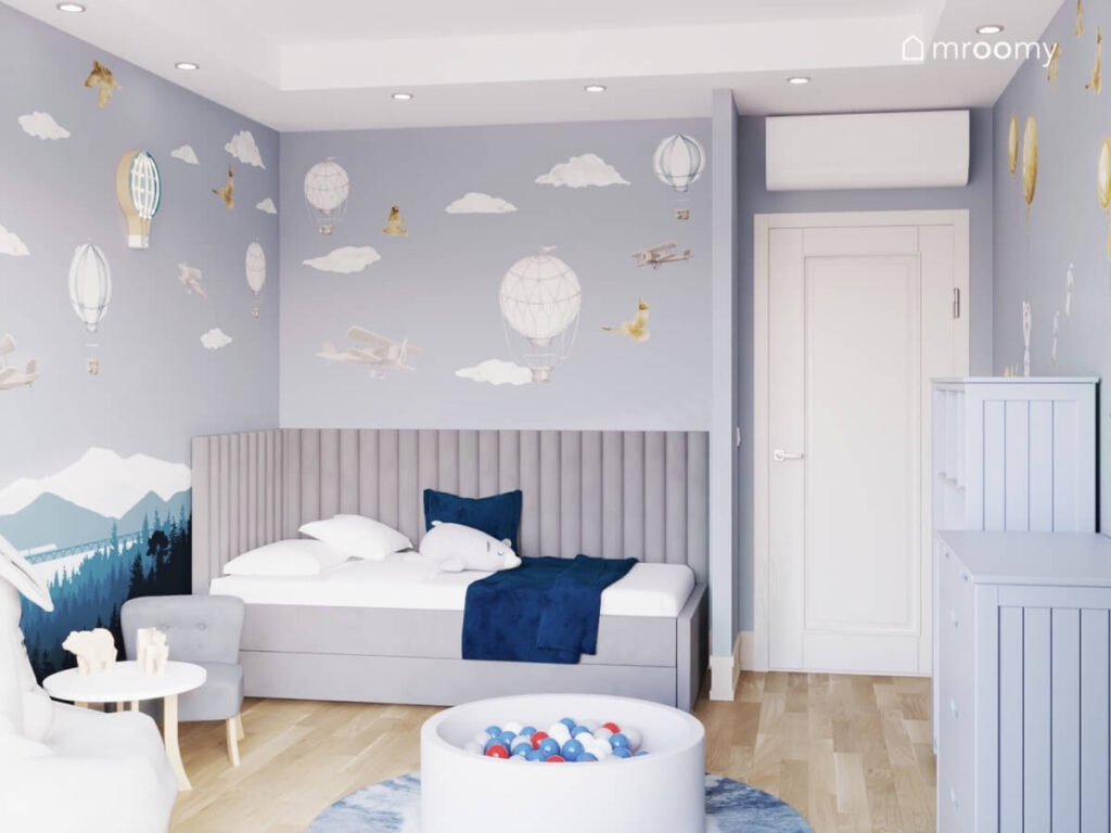 Szare łóżko z miękkimi panelami oraz basen z kulkami w pokoju dla chłopca a na ścianach lampki i naklejki balony oraz chmurki
