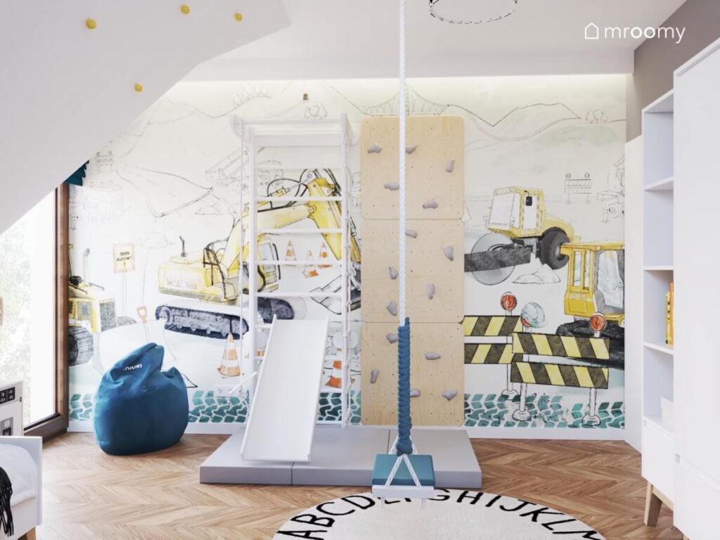 Ściana w pokoju chłopca pokryta wielkoformatową tapetą w pojazdy budowlane a na niej drabinka gimnastyczna ze zjeżdżalnią oraz ścianka wspinaczkowa