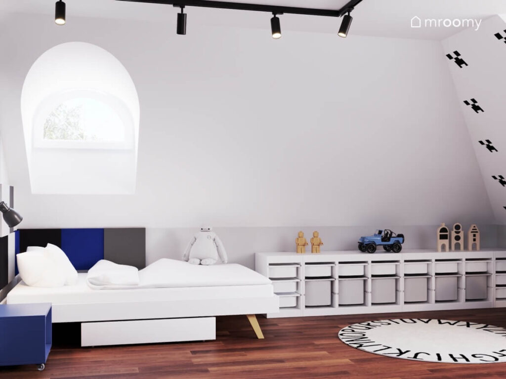 Strefa spania w białym pokoju chłopca w niej proste łóżko uzupełnione kwadratowymi panelami a obok niski regał z pojemnikami i dywan z alfabetem
