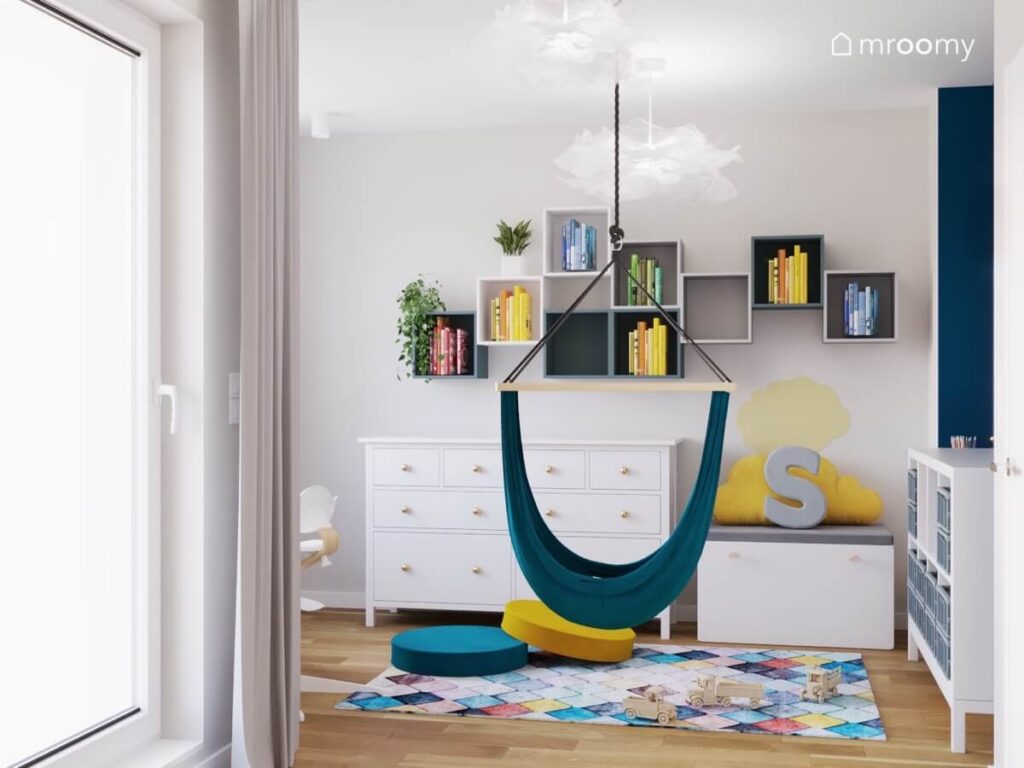 Jasny pokój dla małego chłopca z półkami ściennymi oraz białą komodą a także kolorowym dywanem i pufkami na podłodze