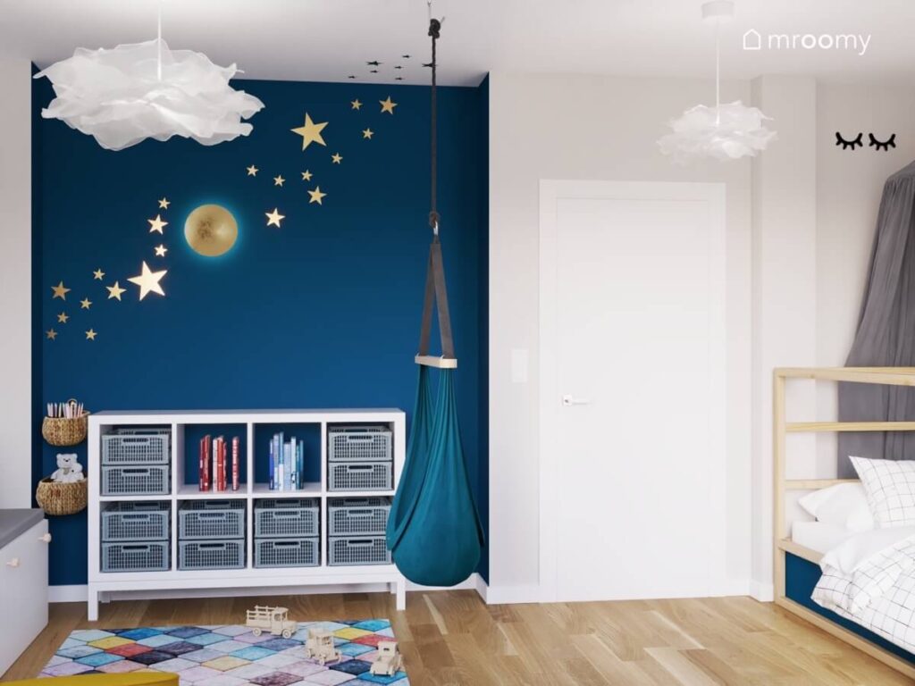 Biało niebieski pokój dla małego chłopca z niskim regałem huśtawką wiszącą oraz złotymi gwiazdkami i lampką księżyc na ścianie