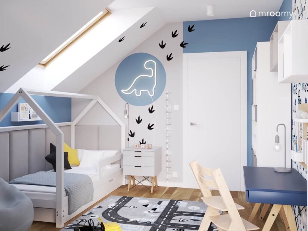 Szaro niebieski poddaszowy pokój dla chłopca z białym łóżkiem domkiem dywanem z drogą oraz ledonem w kształcie dinozaura i śladami łap na ścianach
