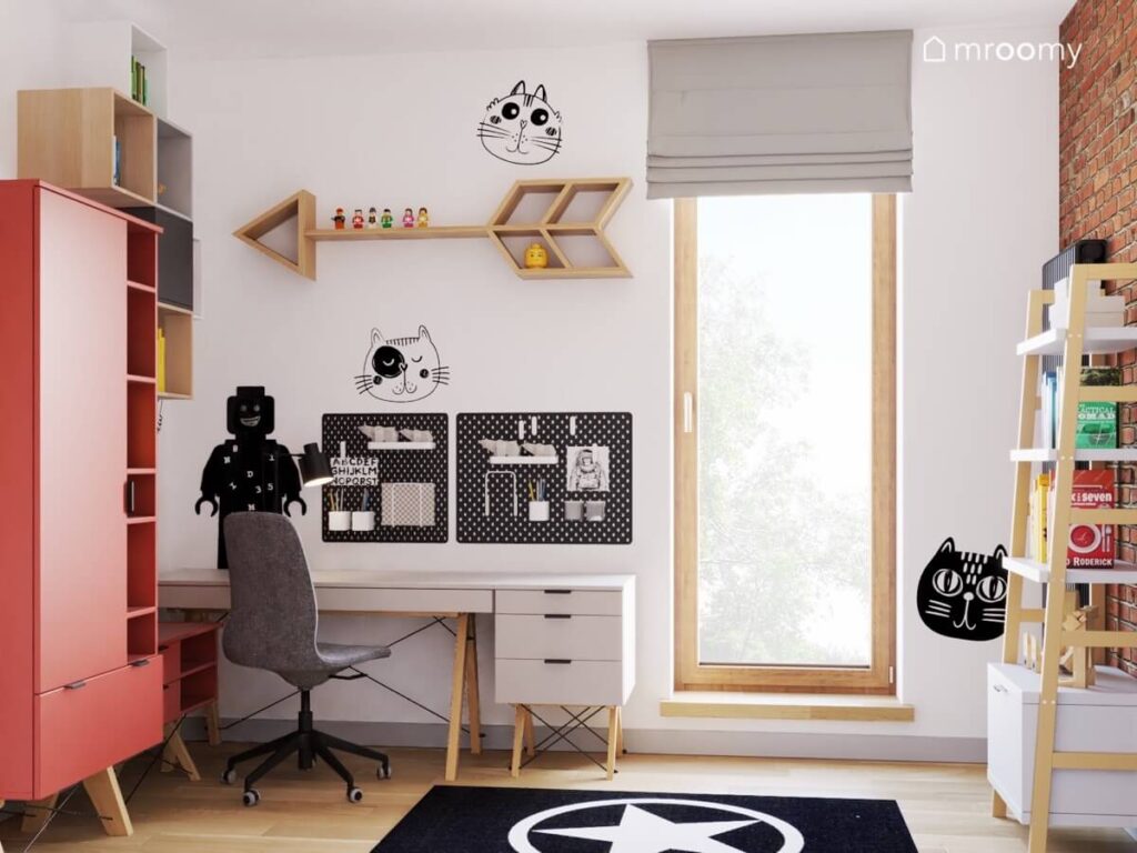 Strefa nauki w jasnym pokoju chłopca a w niej szare biurko z kontenerkiem organizerami i tablicą kredową w kształcie ludzika Lego a powyżej zabawne naklejki w koty i półka w kształcie strzały