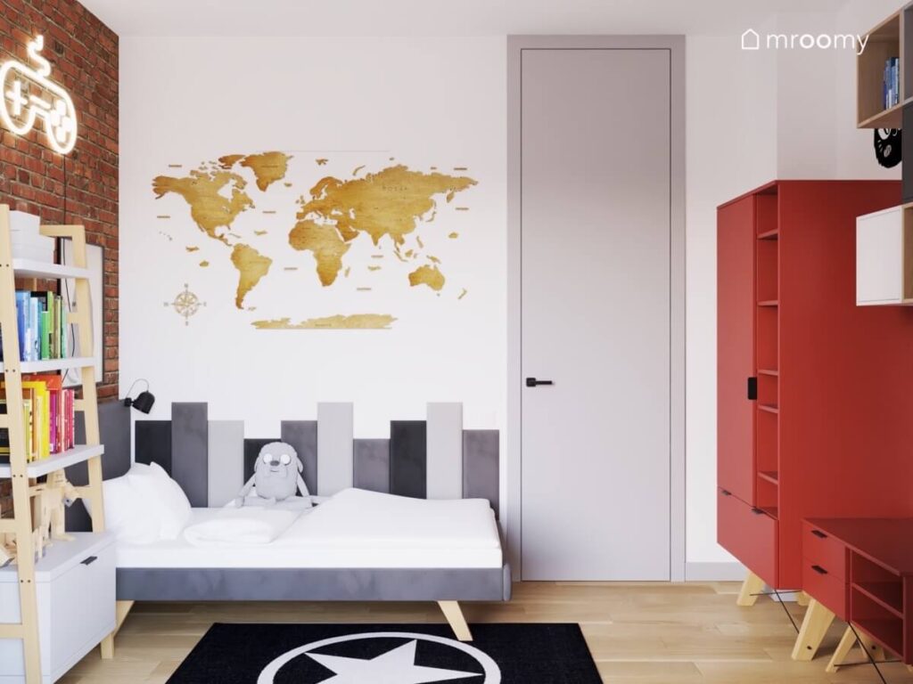 Szare tapicerowane łóżko w pokoju chłopca uzupełnione prostokątnymi panelami w różnych odcieniach szarości oraz drewniana mapa świata a pod drugą ścianą czerwona szafa z półkami