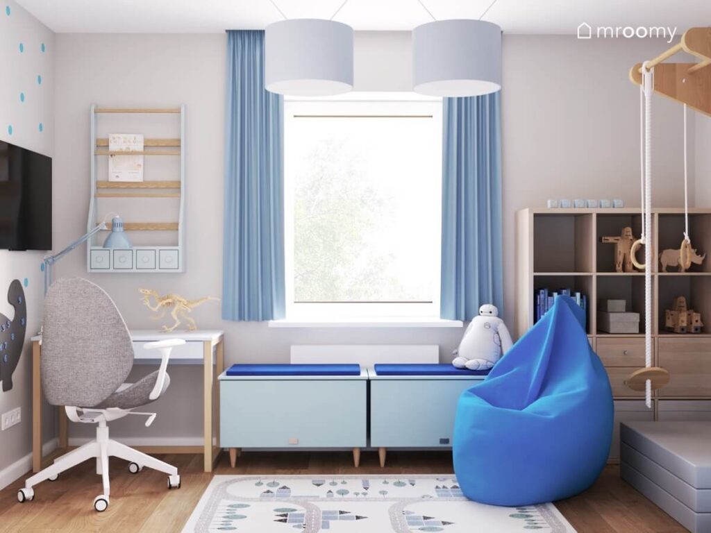 Białe biurko na drewnianych nogach nad nim biblioteczka a obok dwie skrzynie z siedziskami i duża niebieska pufa a na podłodze dywan z miastem w pokoju chłopca