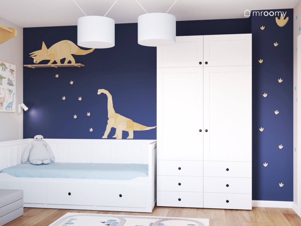 Granatowa ściana w pokoju dla chłopca a na niej białe łóżko i szafa oraz sklejkowe dinozaury oraz ich ślady