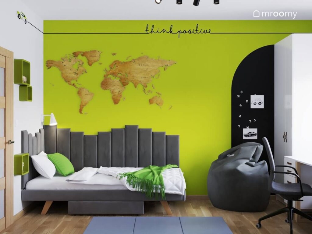 Strefa spania w biało zielonym pokoju chłopca a w niej szare łóżko uzupełnione szarymi panelami ściennymi oraz drewniana mapa świata na ścianie a obok pufa i powierzchnia magnetyczna