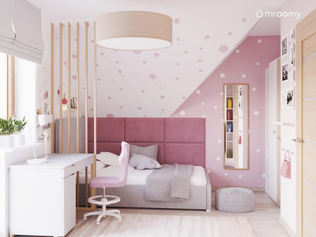 Strefa spania w różowo białym pokoju dziewczynki a w niej szare łóżko uzupełnione panelami ściennymi obok prostokątne lustro oraz biurko a na suficie duża lampa