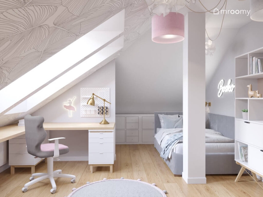 Biało szary poddaszowy pokój dla nastolatki a w nim szare tapicerowane łóżko białe biurko z drewnianym blatem oraz tapeta w liście na skosie