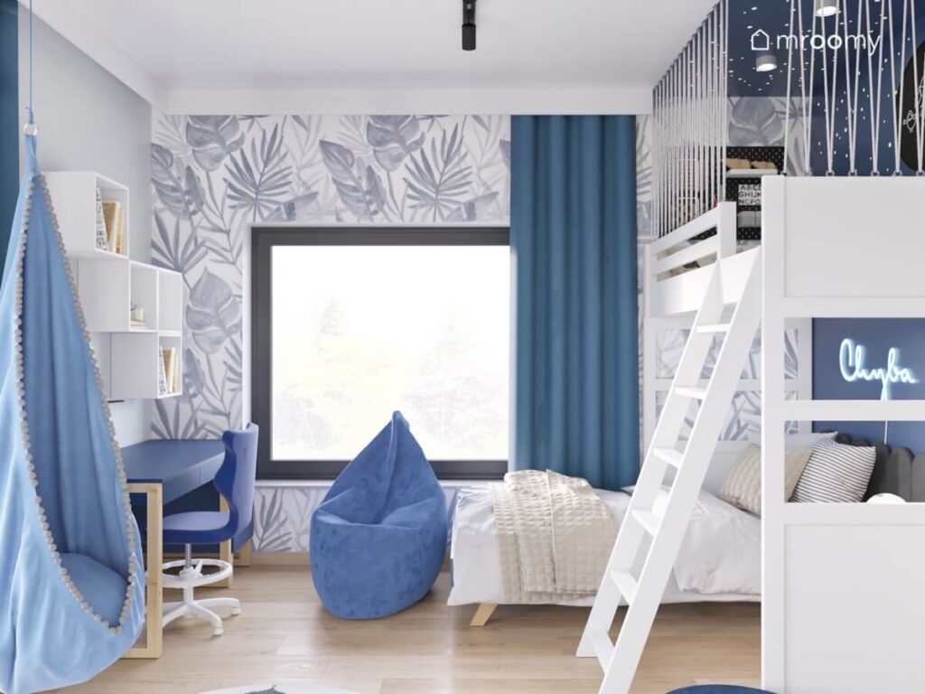 Ściana pokryta tapetą w liście a pod nią łóżko oraz niebieska pufa w pokoju dla dziewczynki z białą antresolą