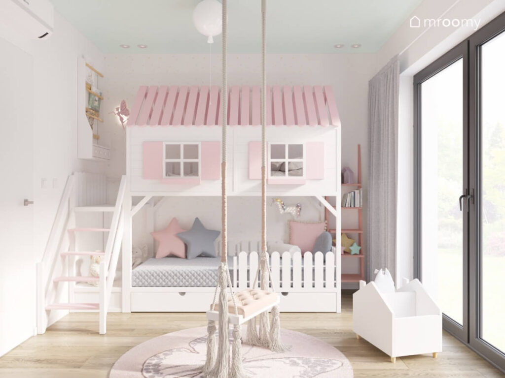 Biało różowa antresola w formie domku z wygodnym miejscem do siedzenia pod spodem a na środku huśtawka z ozdobnymi frędzlami dywan z motylem i biblioteczka w pokoju dziewczynki