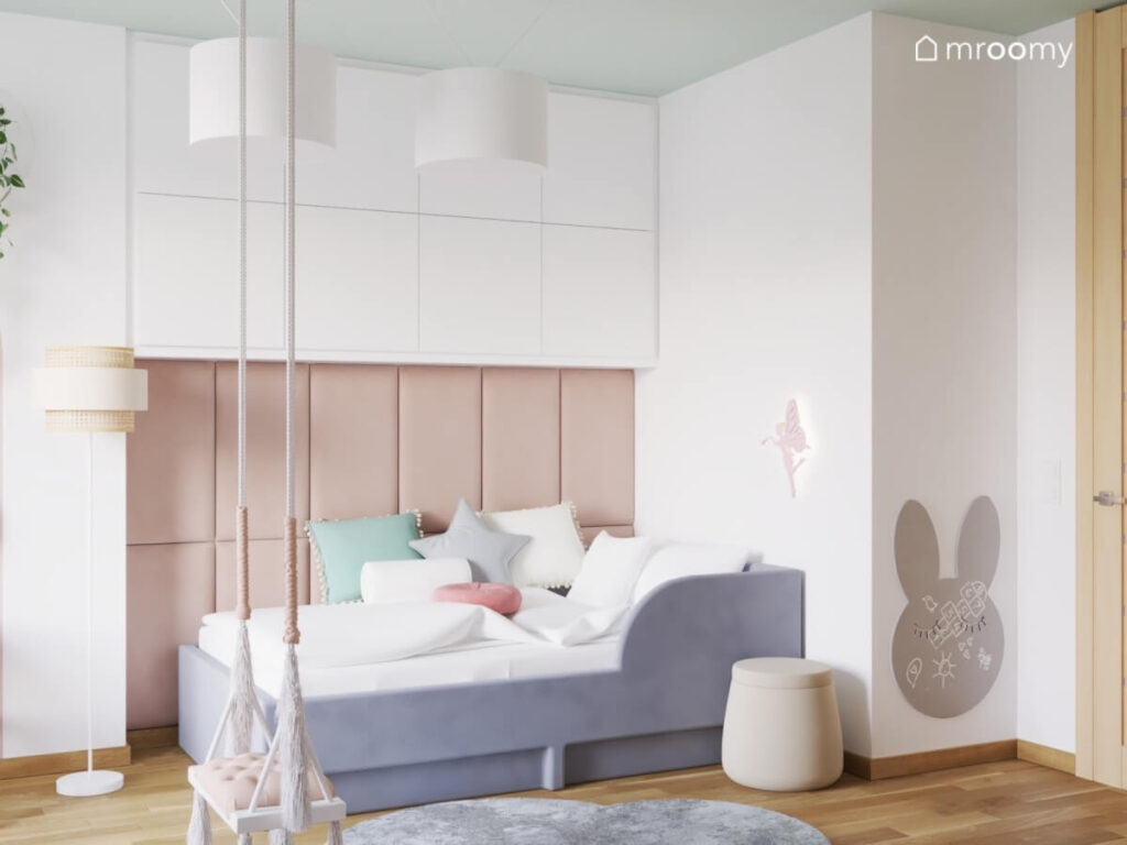 Strefa spania w pokoju dziewczynki a w niej tapicerowane łóżko z dużą liczbą poduszek oraz lampka wróżka