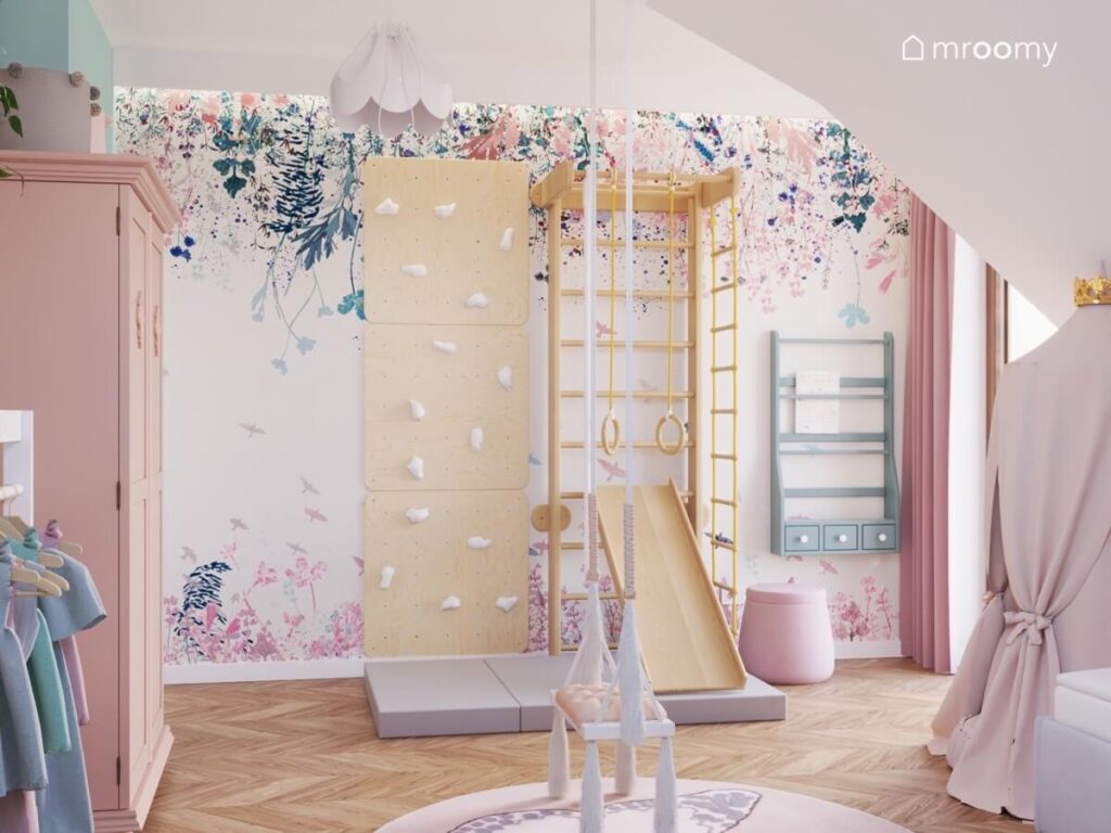 Ściana pokryta tapetą w kwiaty a na niej drewniany zestaw gimnastyczny ze ścianką wspinaczkową i drabinką a obok niebieska biblioteczka i różowa pufa w pokoju dziewczynki