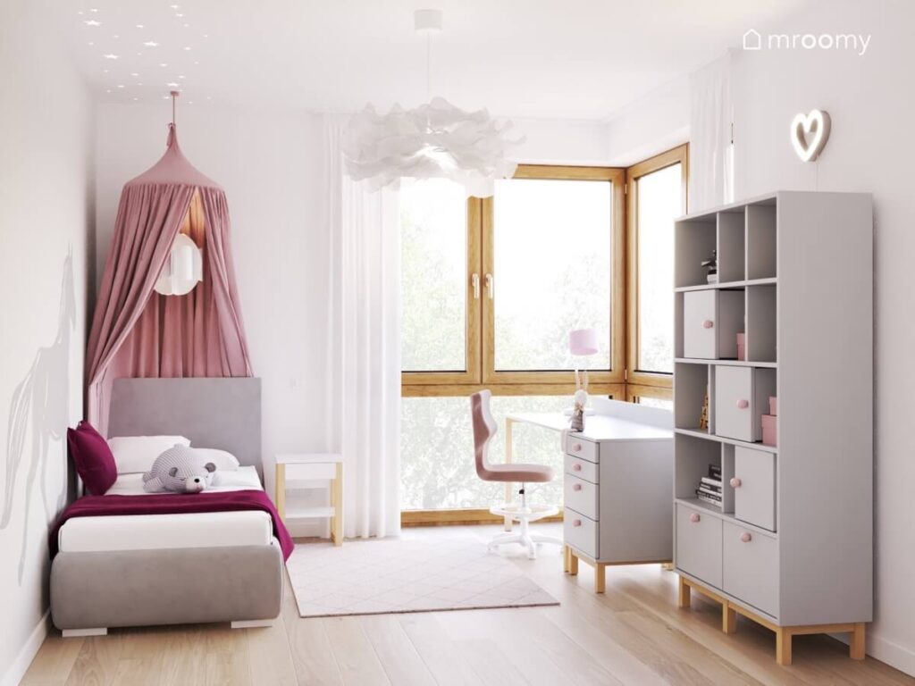 Jasny pokój dla dziewczynki a w nim tapicerowane łóżko z baldachimem oraz szary regał i białe biurko z kontenerkiem