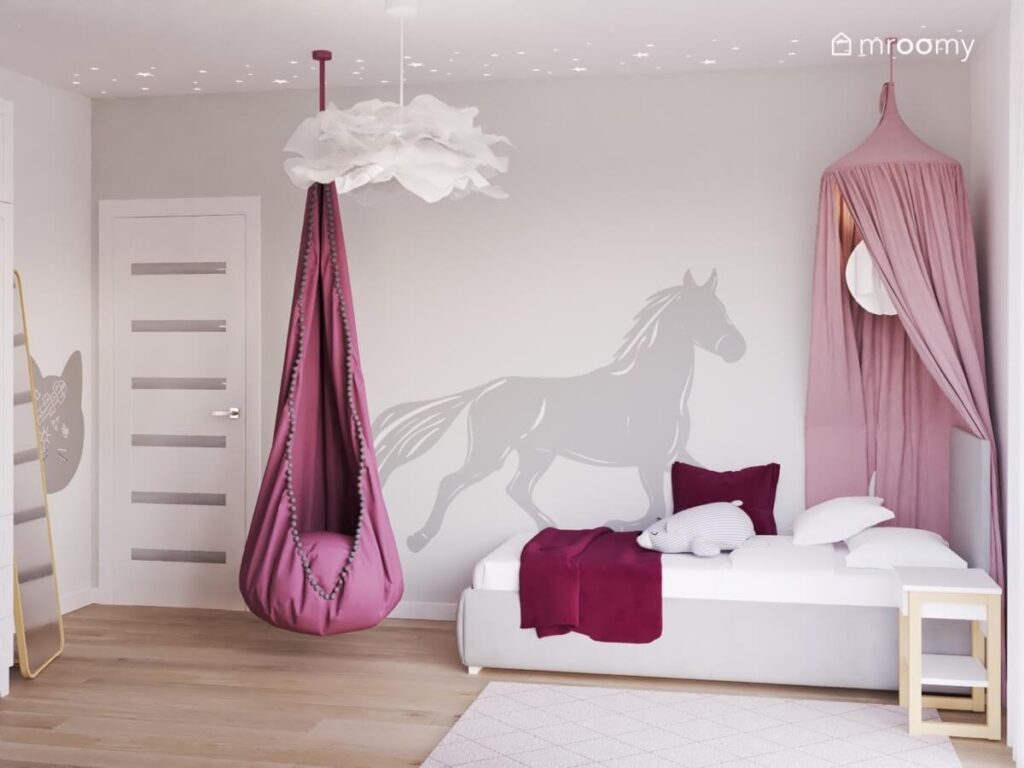 Strefa spania w pokoju dla dziewczynki a w niej szare tapicerowane łóżko z różowym baldachimem oraz naklejka koń a na środku fotel kokon oraz papierowa lampa sufitowa