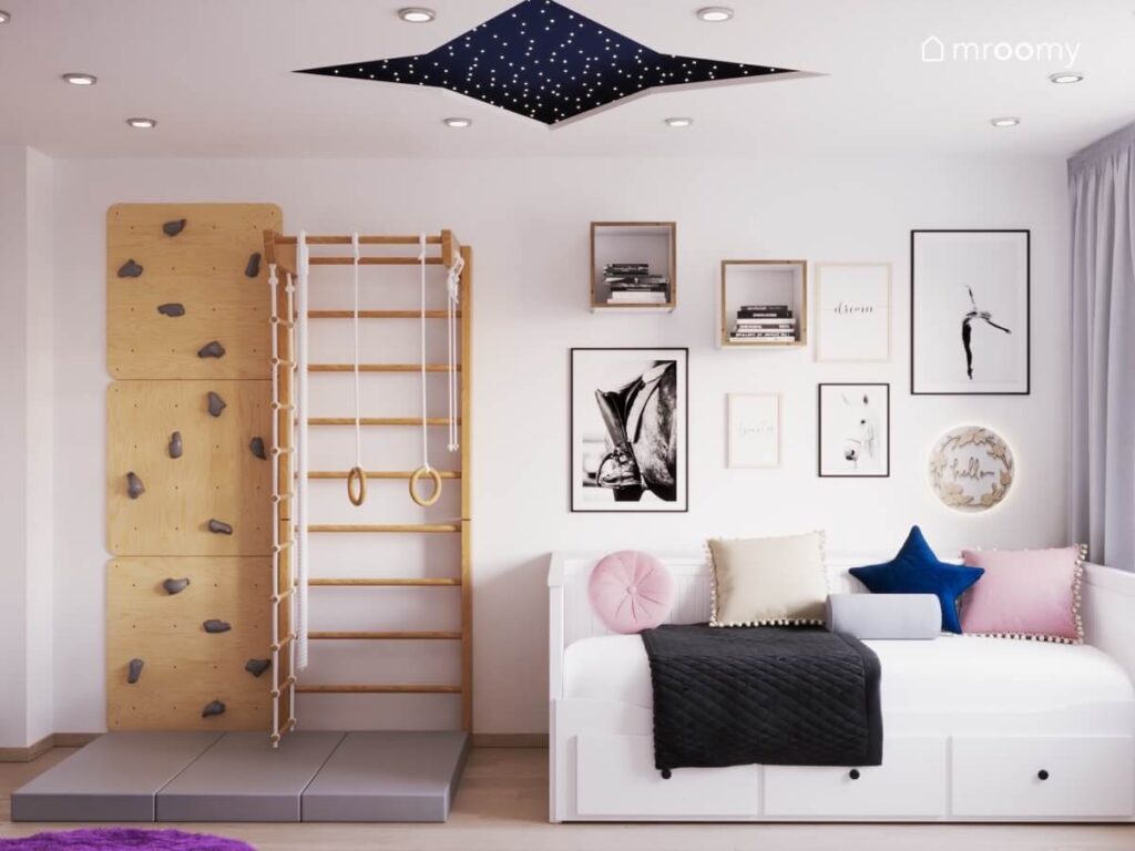 Drewniane panele wspinaczkowe i drabinka gimnastyczna a obok łóżko i ozdobne plakaty w białym pokoju dziewczynki