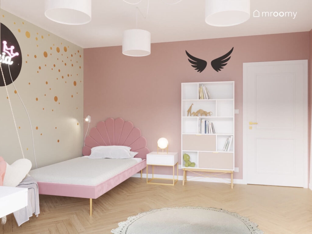 Różowo beżowy pokój dla dziewczynki a w nim różowe łóżko z ozdobnym zagłówkiem biały regał oraz złote kropki i angielskie skrzydła na ścianach