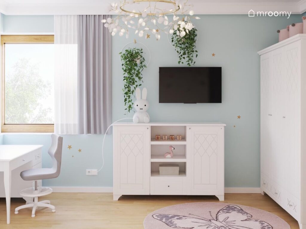 Błękitna ściana w pokoju dla dziewczynki a na niej telewizor biała szafka oraz dwa kwietniki