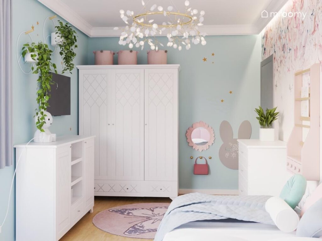 Duża biała szafa z ozdobnymi frontami a obok małe lusterko i tablica kredowa królik a na suficie ozdobna lampa w błękitnym pokoju dziewczynki