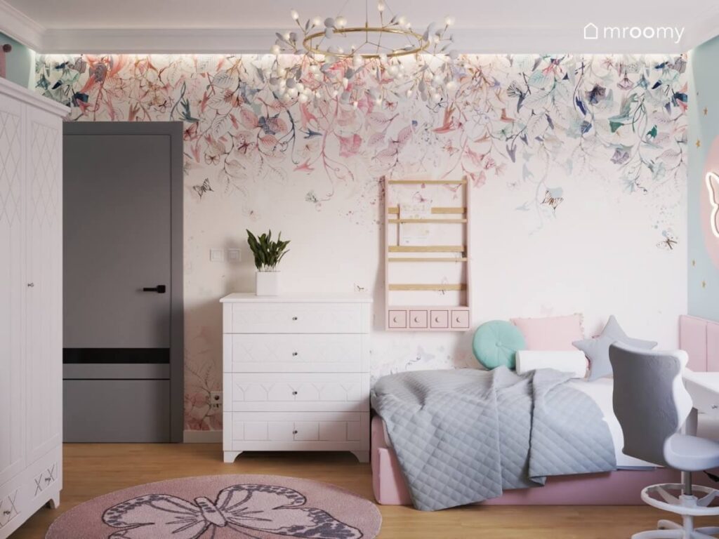 Ściana pokryta tapetą w kwiaty na niej biblioteczka szafka oraz różowe łóżko a na suficie ozdobna lampa