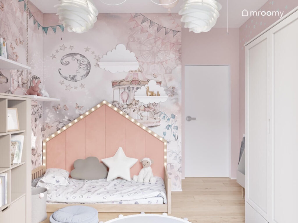 Strefa spania w pokoju małej dziewczynki z drewnianym łóżkiem domkiem uzupełnionym miękkimi panelami i oświetleniem LED a na ścianie bajkowa tapeta
