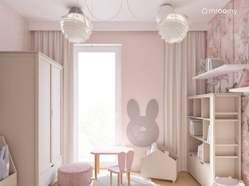 Różowy pokój dla rocznej dziewczynki a w nim beżowa szafa i regał tablica kredowa w kształcie królika oraz ozdobne oryginalne lampy sufitowe