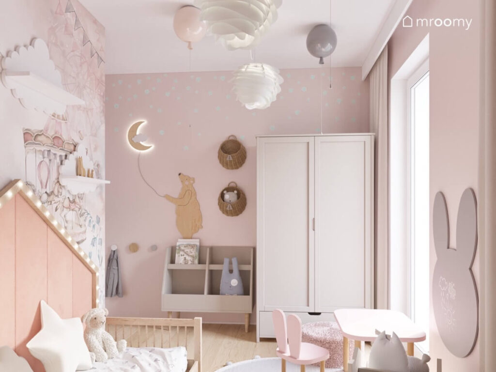 Beżowa szafa biblioteczka kosze na ścianach oraz ozdoba w kształcie niedźwiedzia a u sufitu lampy w kształcie balonów w przytulnym pokoju małej dziewczynki