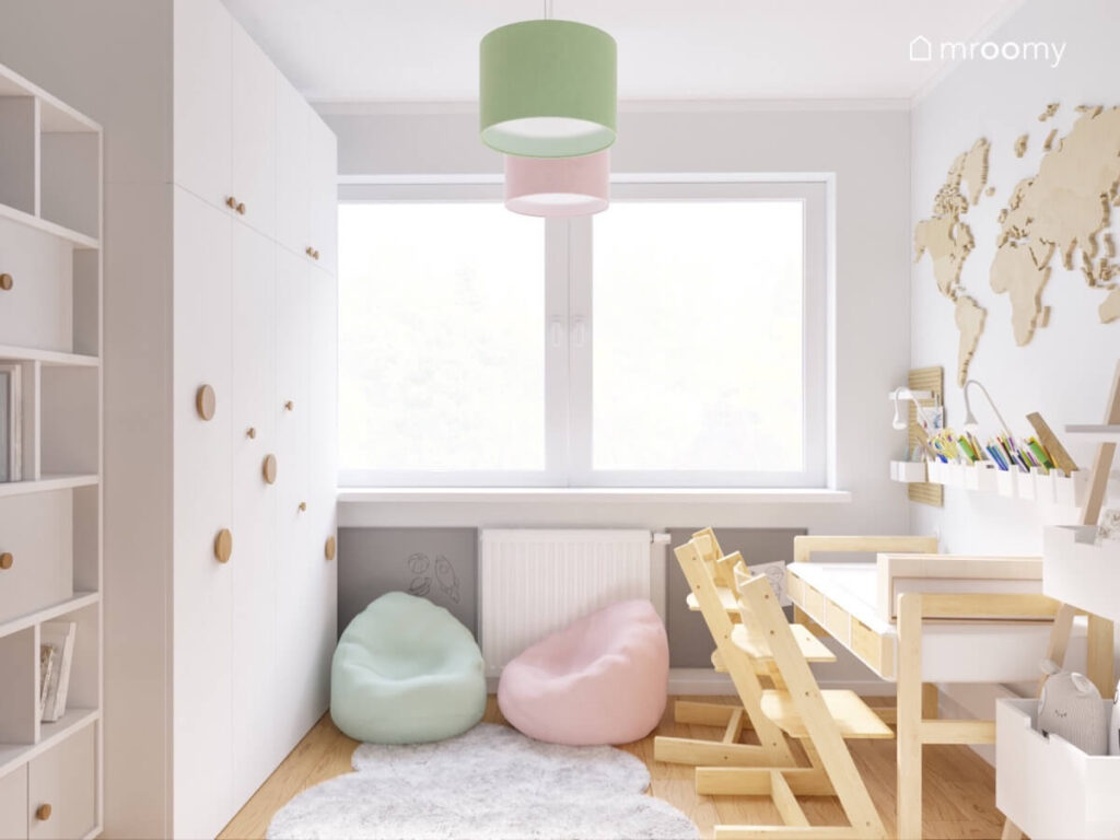 Duża biała szafa z drewnianymi uchwytami naprzeciwko biurko z drewnianymi szufladami i krzesłami a na środku pastelowe pufy i dywan chmurka w pokoju dla rodzeństwa