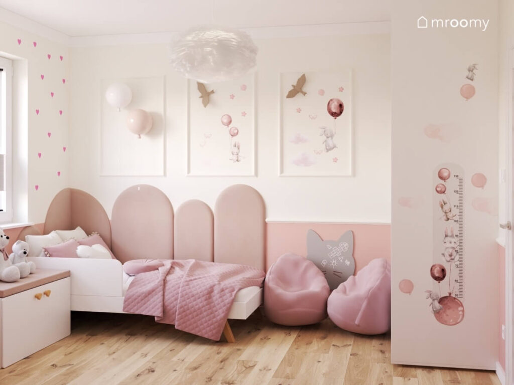 Białe łóżko uzupełnione miękkimi panelami a obok różowe pufy i tablica kredowa kot a na ścianie sztukaterie z ozdobami