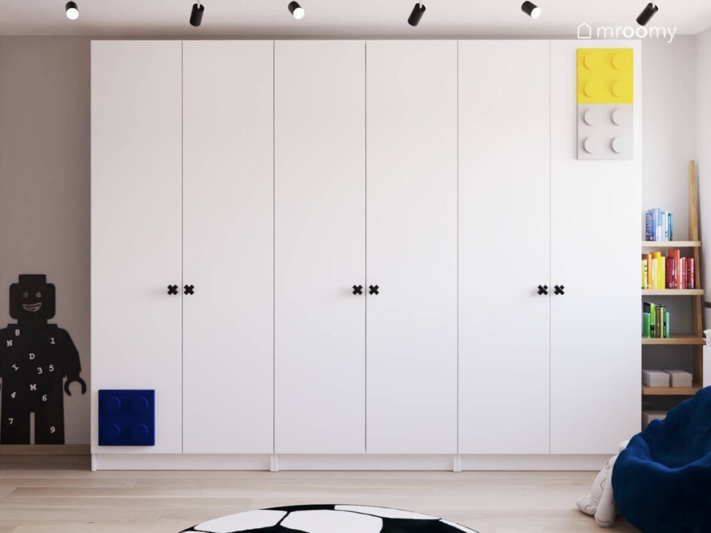 Duża biała szafa w pokoju dla chłopców uzupełniona gałkami w kształcie iksów oraz kolorowymi panelami klocki