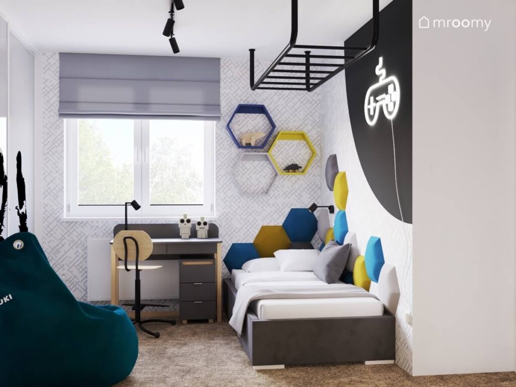 Ściana pokryta tapetą w minimalistyczny wzór na niej biurko oraz kolorowe półki heksagony a poniżej ciemnoszare tapicerowane łóżko i miękkie panele w pokoju chłopca