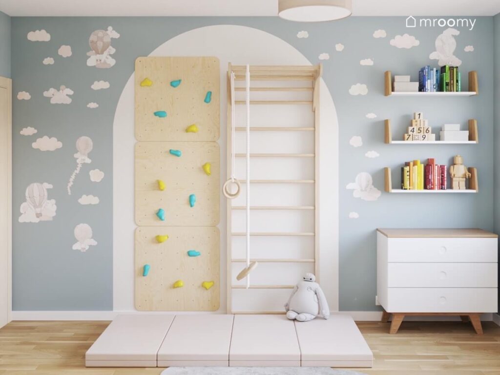 Panele wspinaczkowe ze sklejki z kolorowymi uchwytami oraz drewniana drabinka gimnastyczna na podłodze materac a dookoła naklejki chmurki i balony w biało niebieskim pokoju dla chłopca