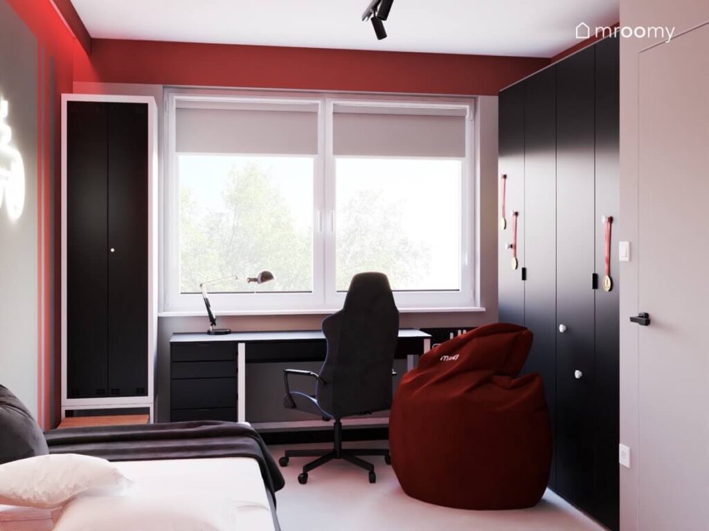 Czarna wąska szafa czarne biurko oraz czarna duża szafa a także czerwona pufa w pokoju dla chłopca wieku szkolnym