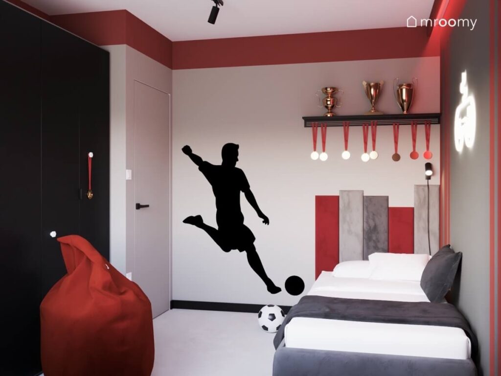 Szaro czerwony pokój dla chłopca w wieku szkolnym a w nim szare tapicerowane łóżko uzupełnione miękkimi panelami naklejka piłkarz czerwona pufa oraz miejsce na puchary i medale