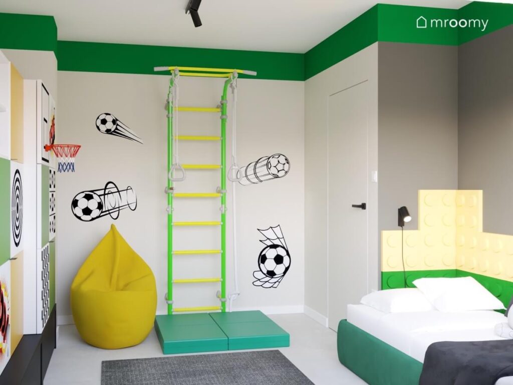 Zielono żółta drabinka gimnastyczna obok niej żółta pufa a na ścianie naklejki w kształcie piłek w szaro zielonym pokoju chłopca