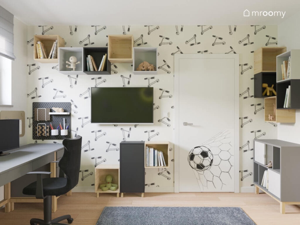 Ściana w pokoju chłopca pokryta tapetą w hulajnogi a na niej kwadratowe szafki telewizor oraz organizer
