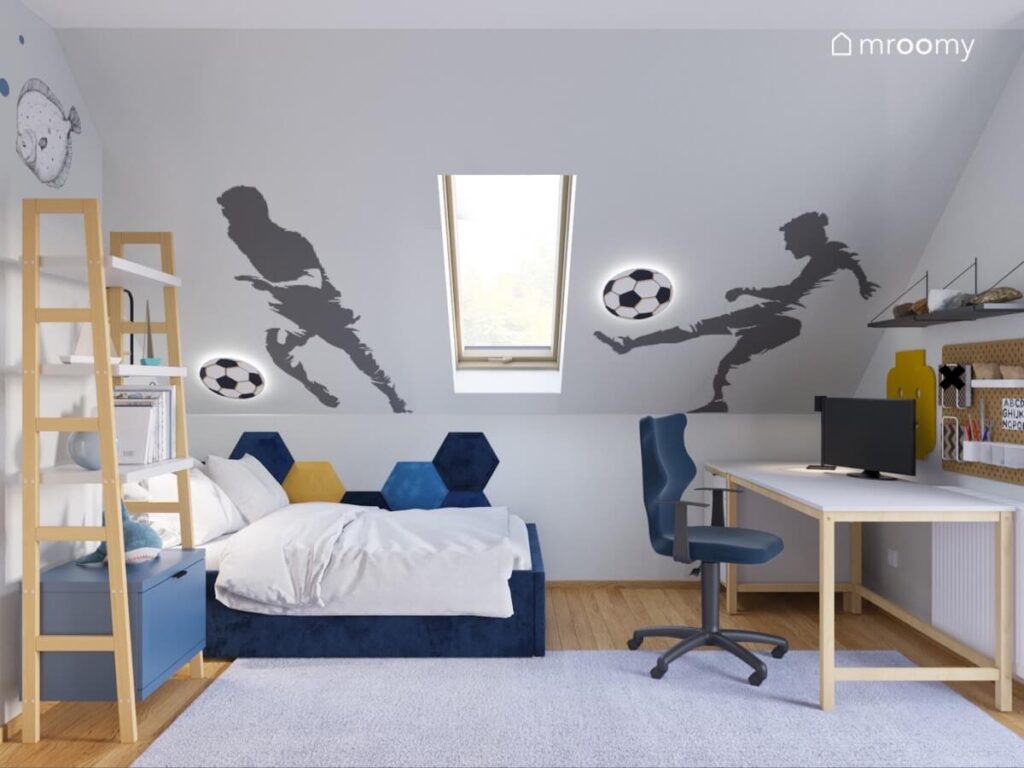 Skos w pokoju chłopca ozdobiony naklejkami w kształcie piłkarzy i lampkami w kształcie piłek a pod nim granatowe tapicerowane łóżko oraz biurko z komputerem