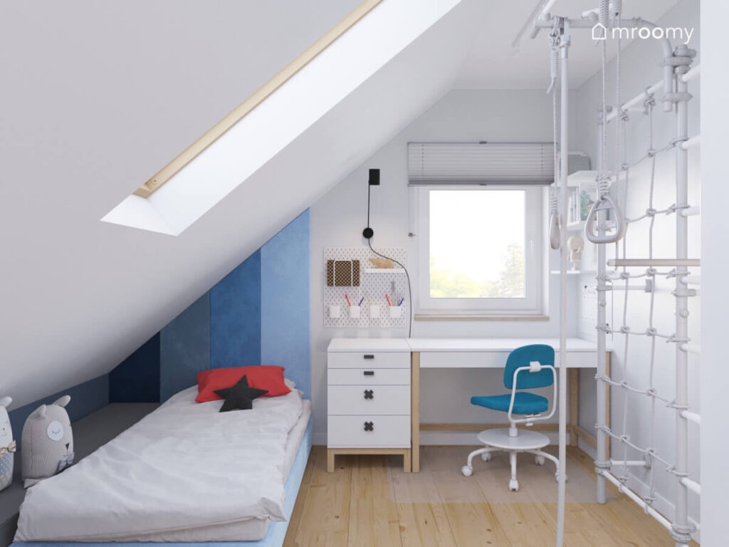 Strefa spania z niebieskim łóżkiem oraz strefa nauki z białym biurkiem na drewnianych nogach w pokoju chłopca na poddaszu