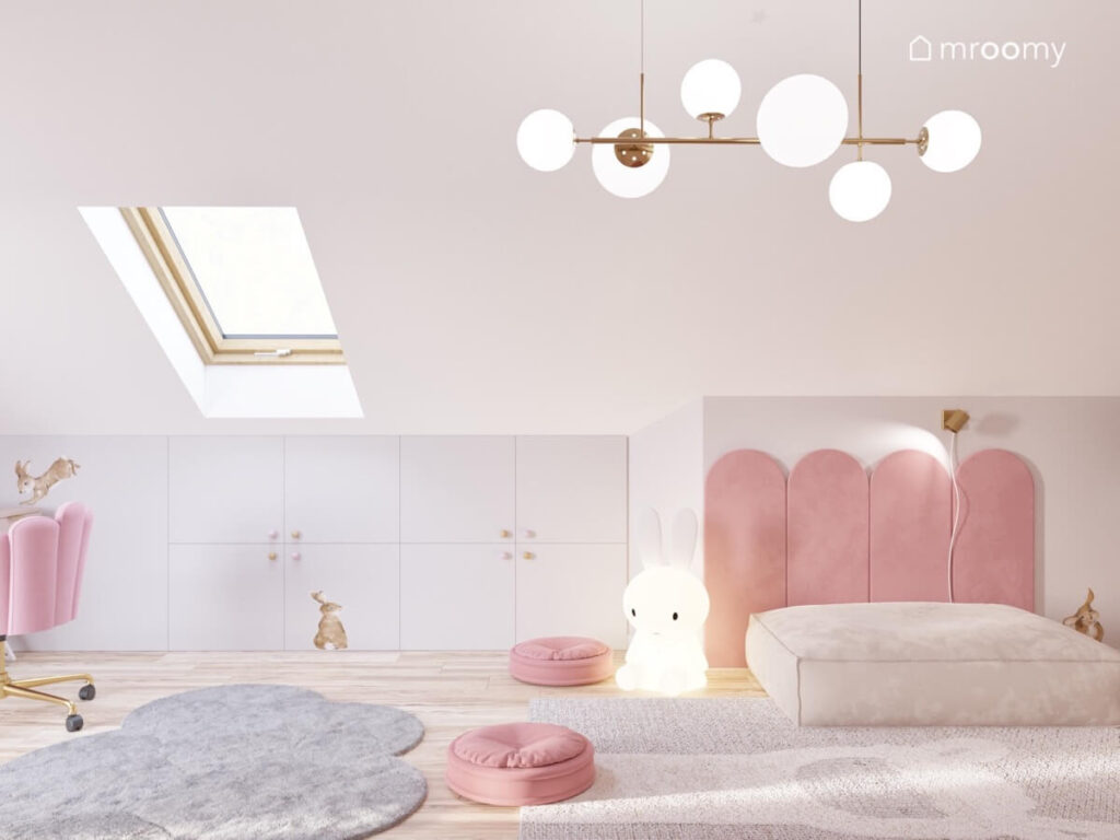 Jasny poddaszowy pokój dla dziewczynki a w nim szare szafki pufy różowe panele ścienne dywan chmurka i mosiężna lampa