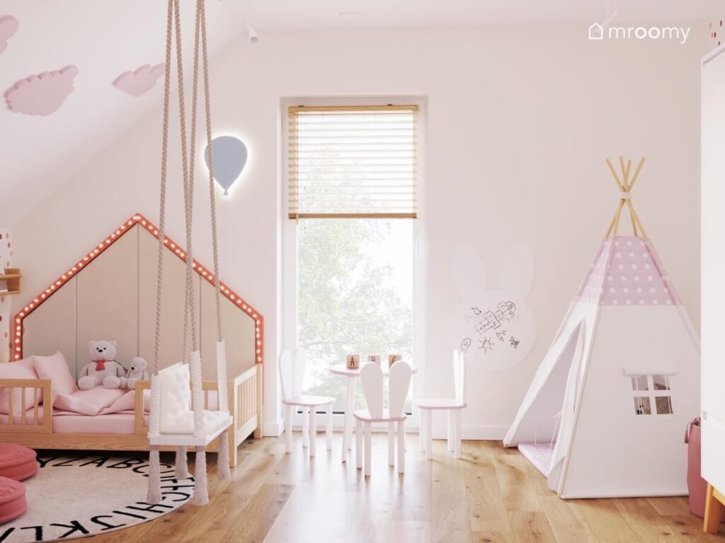 Łóżko domek z ozdobnymi światełkami nad nim kinkiet balon a na środku huśtawka wisząca stolik z krzesełkami oraz namiot tipi w pokoju malutkiej dziewczynki