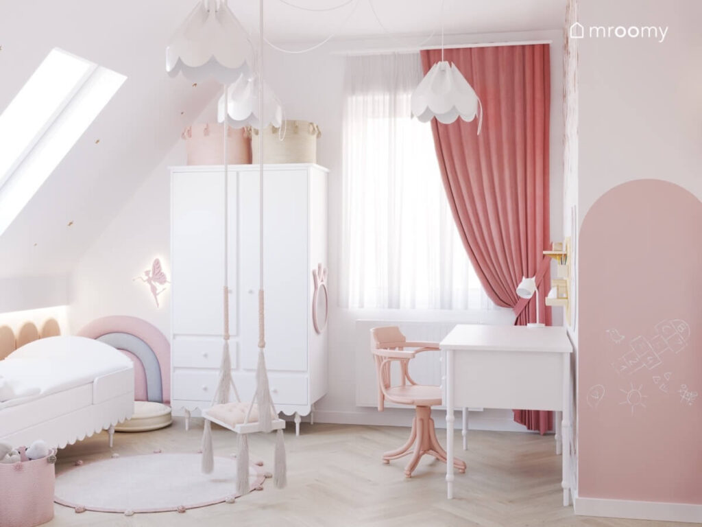 Jasny poddaszowy pokój dla dziewczynki a w nim białe meble różowe dodatki huśtawka wisząca z frędzlami oraz lampy bezy