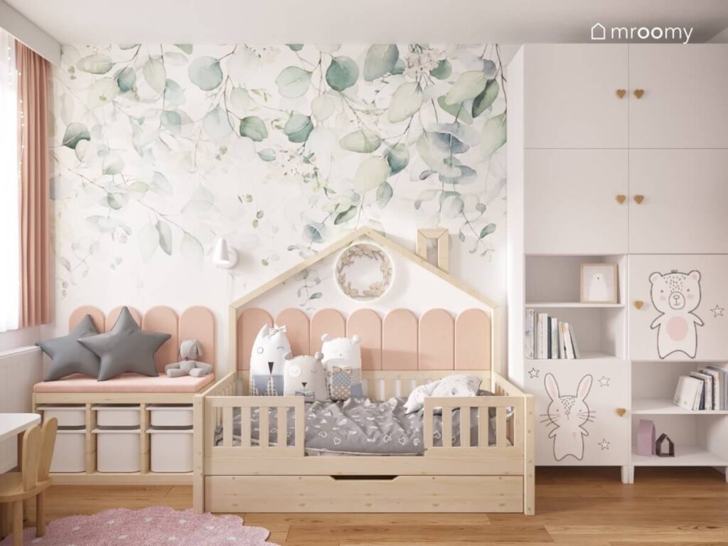 Strefa spania w pokoju malutkiej dziewczynki a w niej drewniane łóżko domek obok regał z pojemnikami i duża biała szafa a na ścianie tapeta w liście
