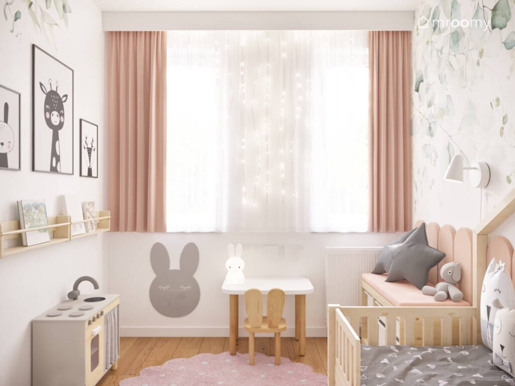 Okno w pokoju małej dziewczynki ozdobione różowymi zasłonami firanką i światełkami a pod spodem tablica kredowa w kształcie królika i stoliczek z krzesłem
