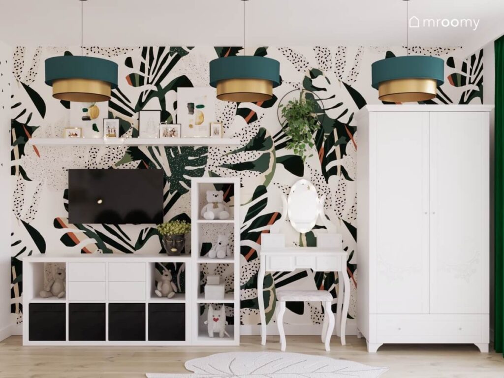 Ściana w pokoju nastolatki pokryta liściastą tapetą a na niej telewizor szafka rtv oraz toaletka a na suficie zielono złote lampy