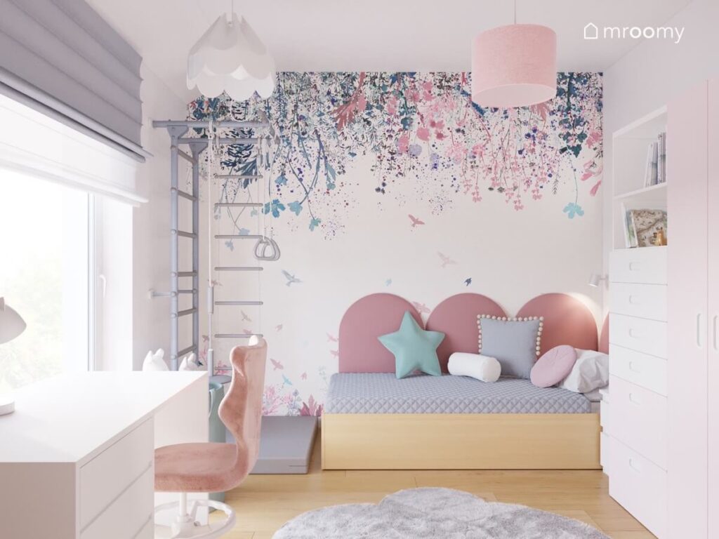 Jasny pokój dla dziewczynki a w nim drewniane łóżko uzupełnione różowymi panelami drabinka gimnastyczna oraz tapeta w kwiaty