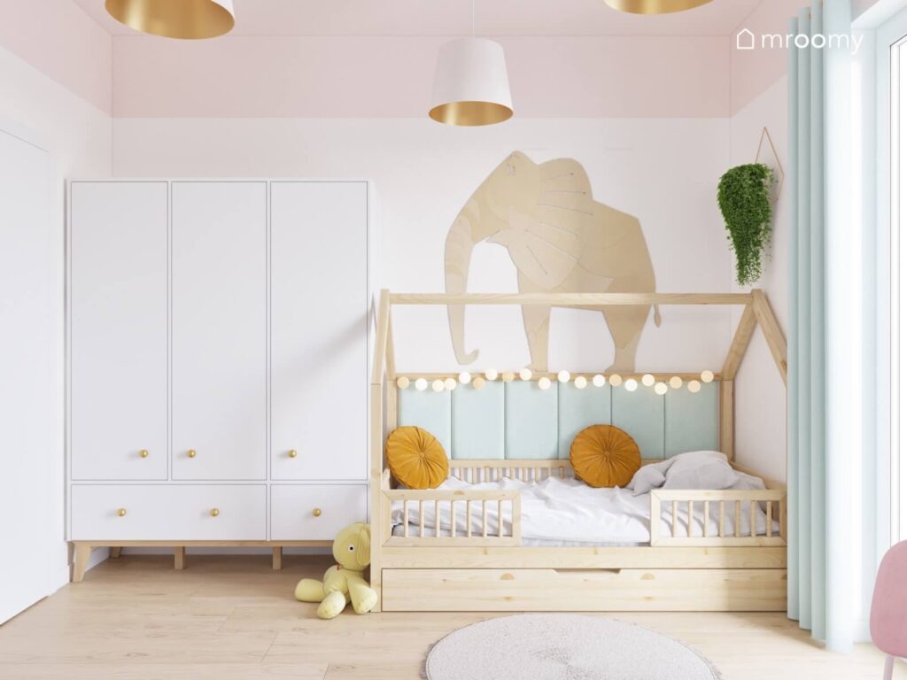 Biała szafa ze złotymi gałkami obok drewniane łóżko domek uzupełnione girlandą cotton balls a na ścianie słoń ze sklejki oraz kwietnik w pokoju dla dziewczynki
