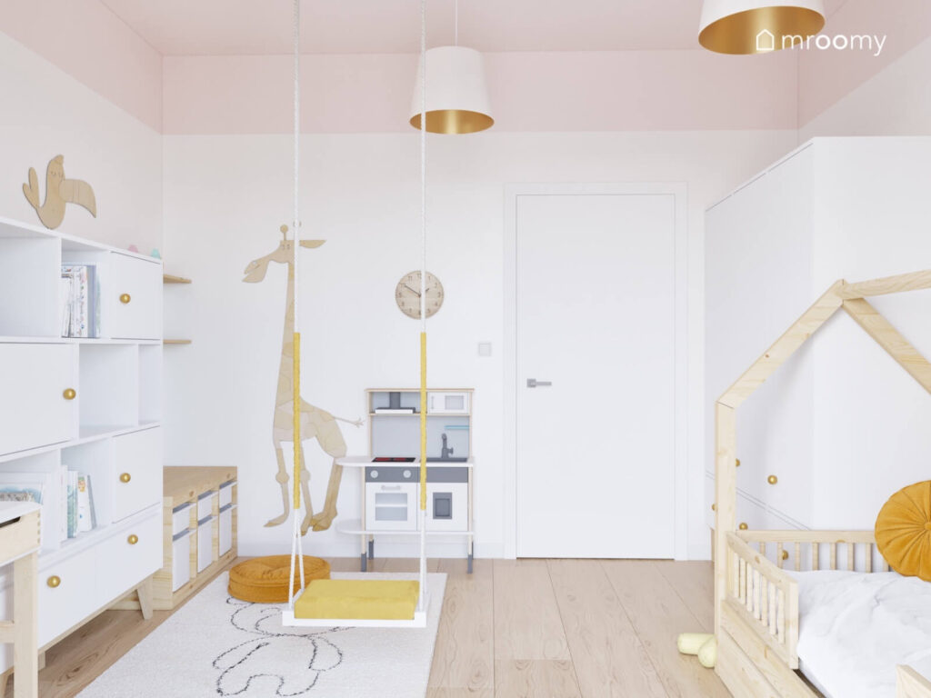 Biały pokój dla małej dziewczynki z różowym sufitem drewnianym łóżkiem domkiem huśtawką wiszącą oraz żyrafą ze sklejki na ścianie