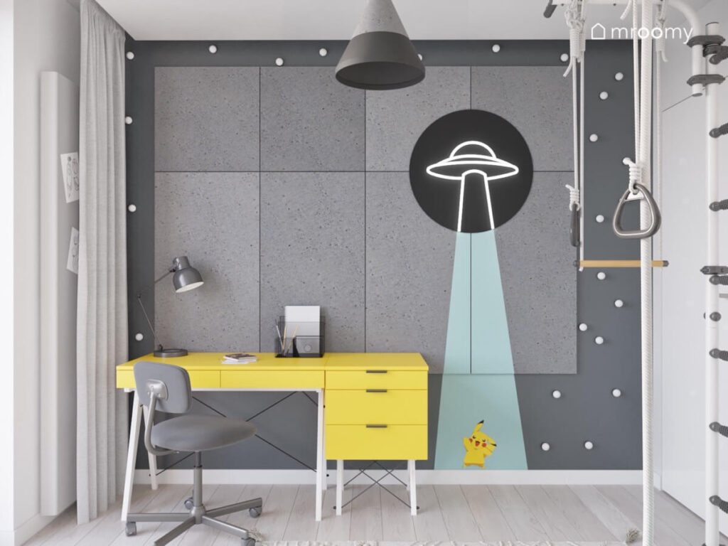 Strefa nauki w pokoju dla chłopca a w niej żółte biurko oraz ściana ozdobiona szarymi panelami białymi gałkami i ledonem w kształcie UFO