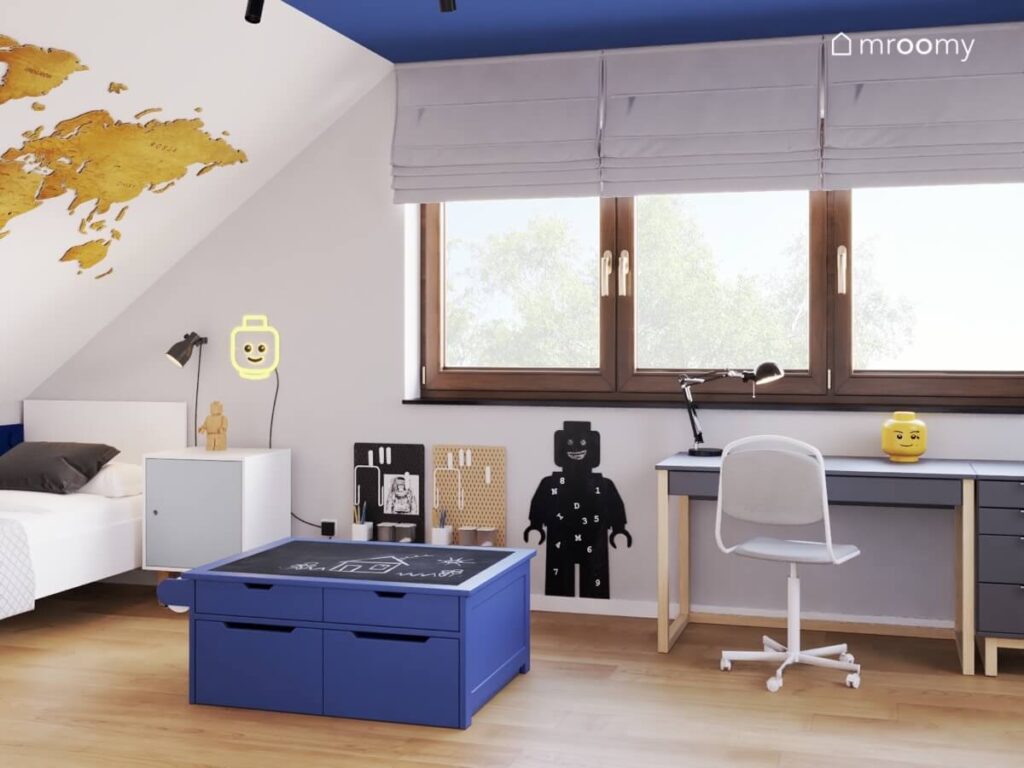 Biało szaro niebieski poddaszowy pokój dla chłopca a w nim mapa świata na skosie stolik do zabawy oraz tablica kredowa w kształcie ludzika Lego